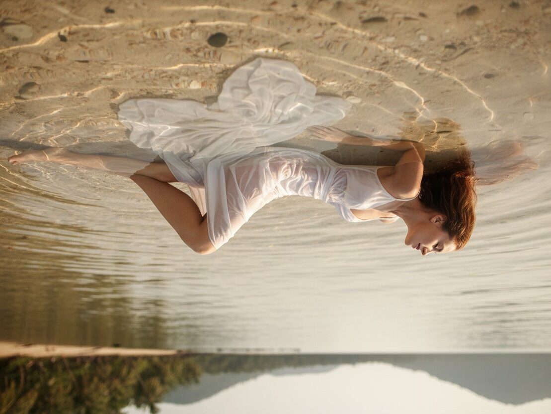 Eine Frau in einem weißen Kleid liegt entspannt im flachen Wasser vor einem tropischen Strand.